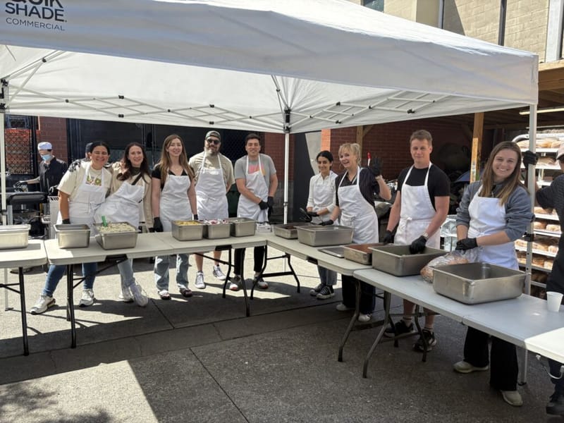 Group of volunteers in white aprons serving food at an outdoor community event under a canopy tent