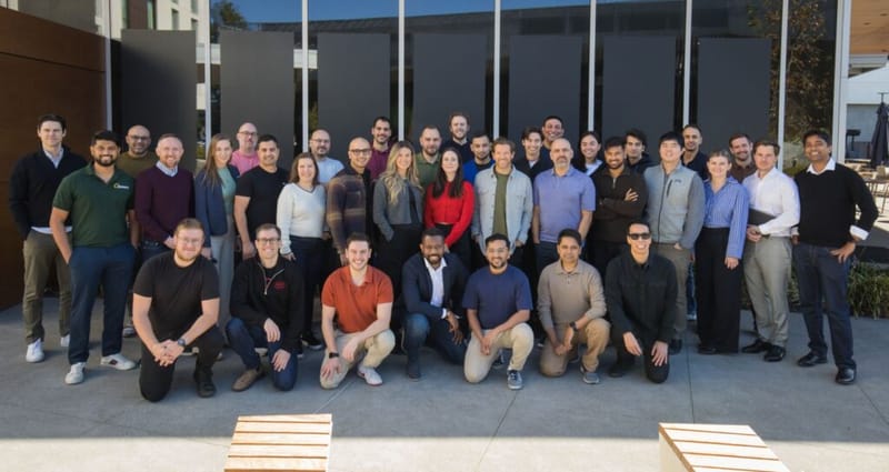 A large group of colleagues posing together outdoors in front of a modern building with dark glass panels