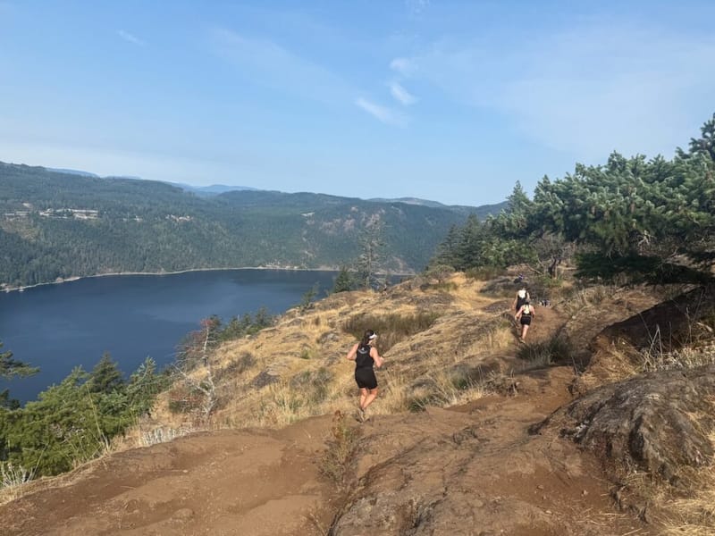 Two trail runners on a rocky ridge above a lake, surrounded by dry grass, conifers, and forested hills