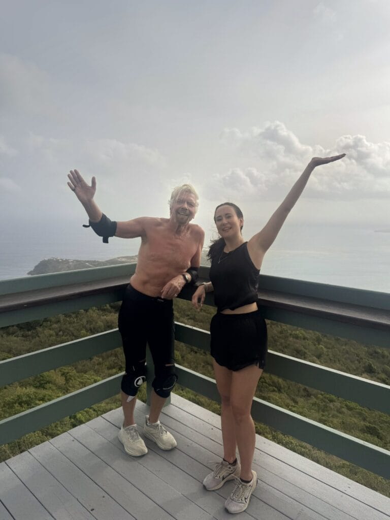 Ania Wysocka and Richard Branson posing with arms raised on a hilltop deck overlooking a tropical coastline