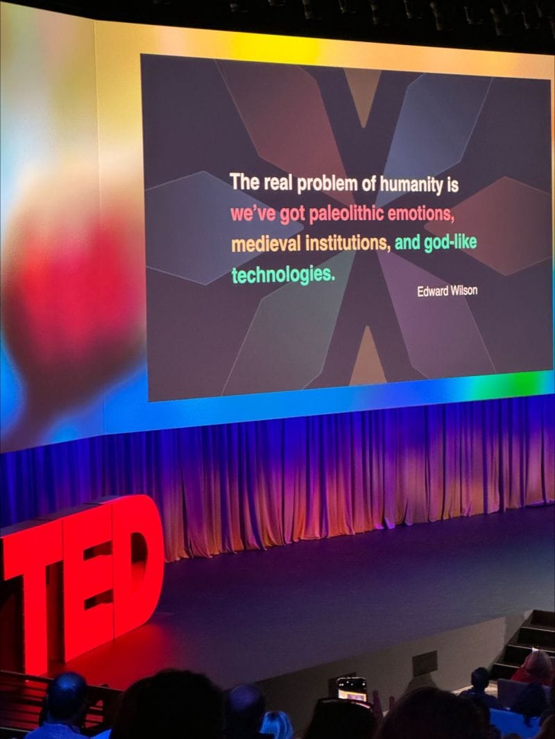 TED conference stage displaying an Edward Wilson quote: "The real problem of humanity is we've got paleolithic emotions, medieval institutions, and god-like technologies." The red TED letters are visible at lower left, with audience members in silhouette below.